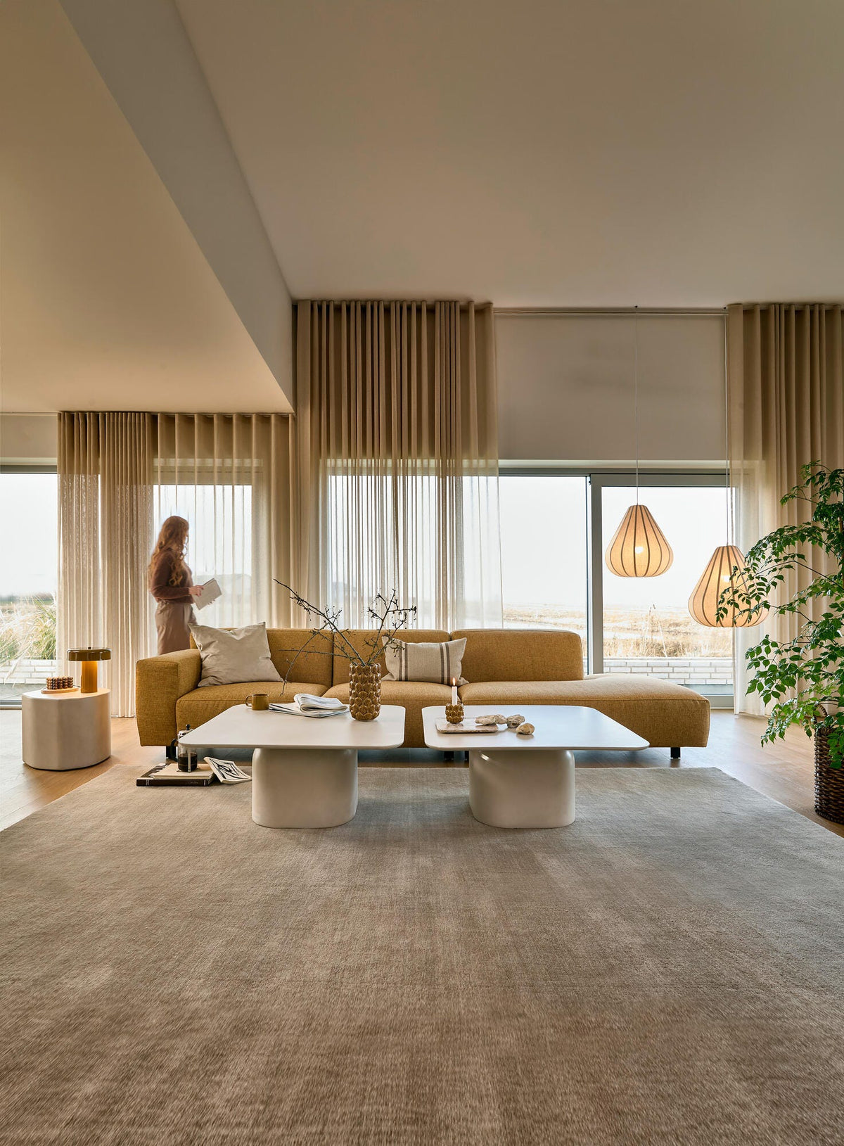 Lyst stue med sennepsgul sofa, beige gardiner, to hvide sofaborde, store vinduer og pendler; en person står ved vinduet.