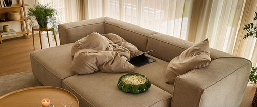 Beige modulsofa med puder og plaid, en laptop og en grøn skål med snacks; rundt sofabord med tændt lys og glas; lyse gardiner, planter og trædetaljer i en lys stue.