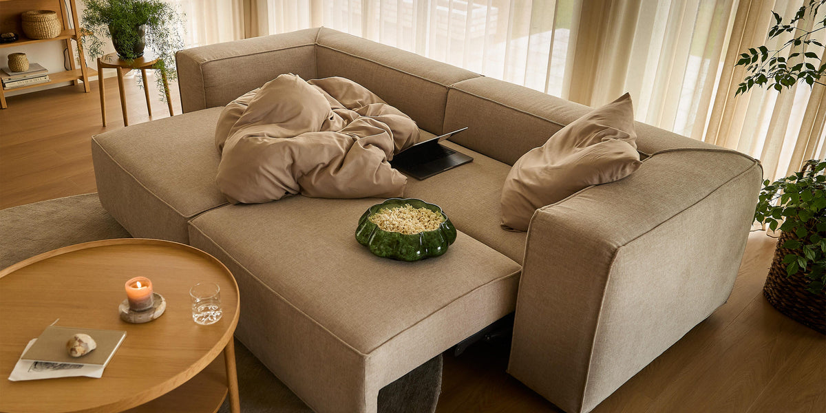 Beige modulsofa med puder og plaid, en laptop og en grøn skål med snacks; rundt sofabord med tændt lys og glas; lyse gardiner, planter og trædetaljer i en lys stue.