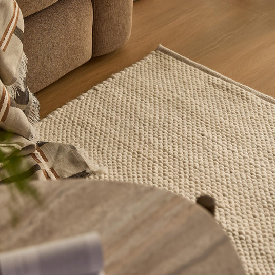 Nærbillede af lyst, tekstureret tæppe i beige ved siden af sofa, plaid og rundt træbord i en varm hjemmeindretning.