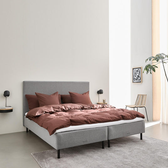 Grå polstret seng med rustfarvet sengetøj i et lyst, minimalistisk soveværelse med væglamper, tæppe, stol og en grøn plante.