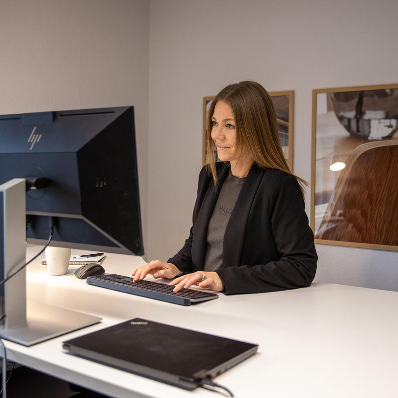 Person arbejder ved et moderne skrivebord med HP-skærm, tastatur og mus, bordlampe og vægkunst; en lukket ThinkPad ligger på bordet, og trøjen har teksten HALO.