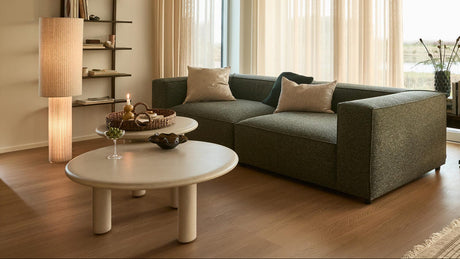Lys, skandinavisk stue med grå modulsofa, lyse runde sofaborde, lette beige gardiner, tændte stearinlys og gulvlampe, hylde med bøger samt konsol med pladespiller og højttalere; tæppe og plante ved store vinduer i dagslys.