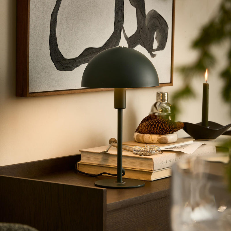 Mørkegrøn bordlampe på bøger med "SOFT MINIMAL", en kogle og et tændt lys på en skænk i en varm, modern indretning med abstrakt vægkunst.
