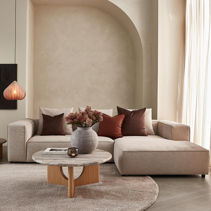 Beige stue med hjørnesofa, rundt sofabord i sten og træ, brune puder, vase med blomster og en hængende lampe i blødt dagslys.