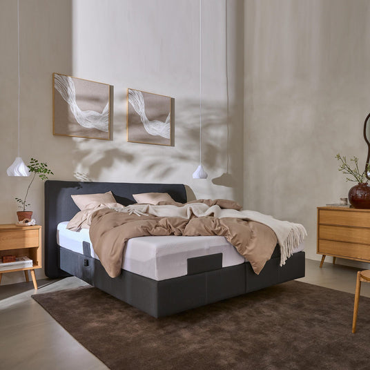Lyst soveværelse med grå polstret seng, beige sengetøj og hvid plaid, to vægbilleder og hvide pendler; TEMPUR ses på madrassen; træmøbler og brun tæppe.