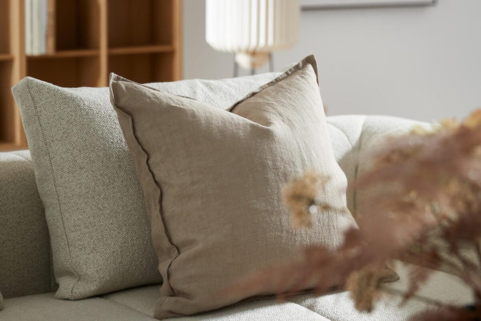 Nærbillede af en beige linnedpude og en lysgrå pude på en lys sofa i stuen; træreol, hvid plisseret bordlampe og tørrede blomster i forgrunden.
