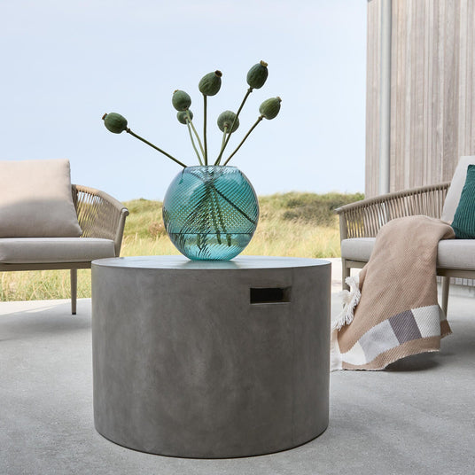 Rundt betonbord på terrasse med turkis glasvase og grønne frøkapsler, loungestole og plaid i baggrunden.