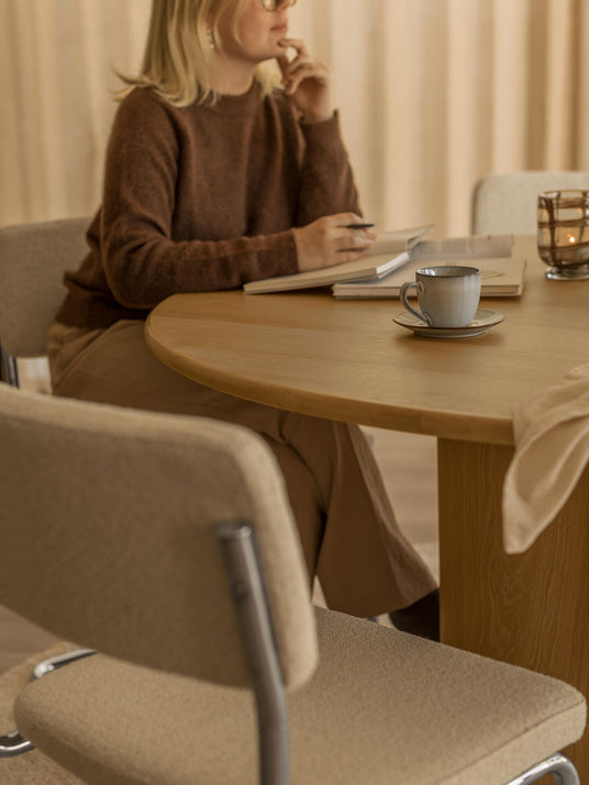 Person sidder ved rundt træbord med notesbog, kop og tændt lyslygte i varm, beige indretning.