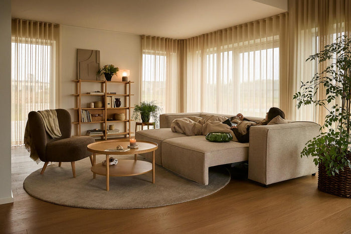 Lyst, varmt stue i nordisk stil med beige modulsofa, rundt tæppe og sofabord i træ; lette beige gardiner, grønne planter og et tændt lys, mens en person slapper af under et tæppe.