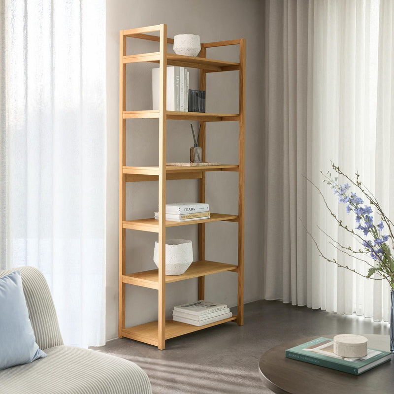 Lyst stueinteriør med åben reol i eg, pyntet med få bøger (bl.a. ”PRADA” og ”New Nordic Houses”) og hvide keramikkar; ved siden af sofa, lette gardiner og et sofabord med vase og blomster.