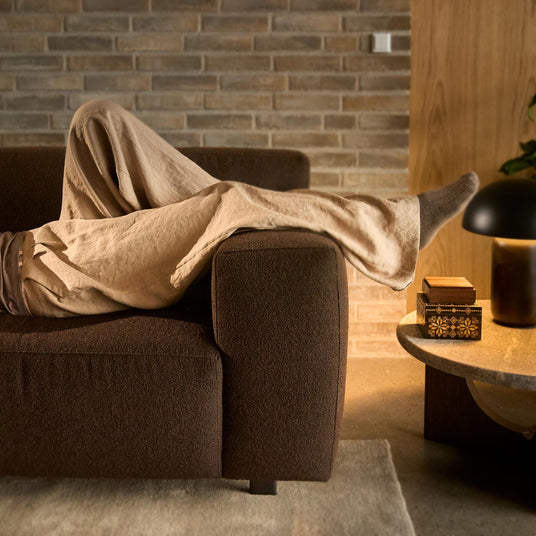 Ben der hviler på en mørkebrun sofa i en hyggelig stue med varm bordlampe, murstensvæg og en lille dekorativ æske på et rundt sofabord.