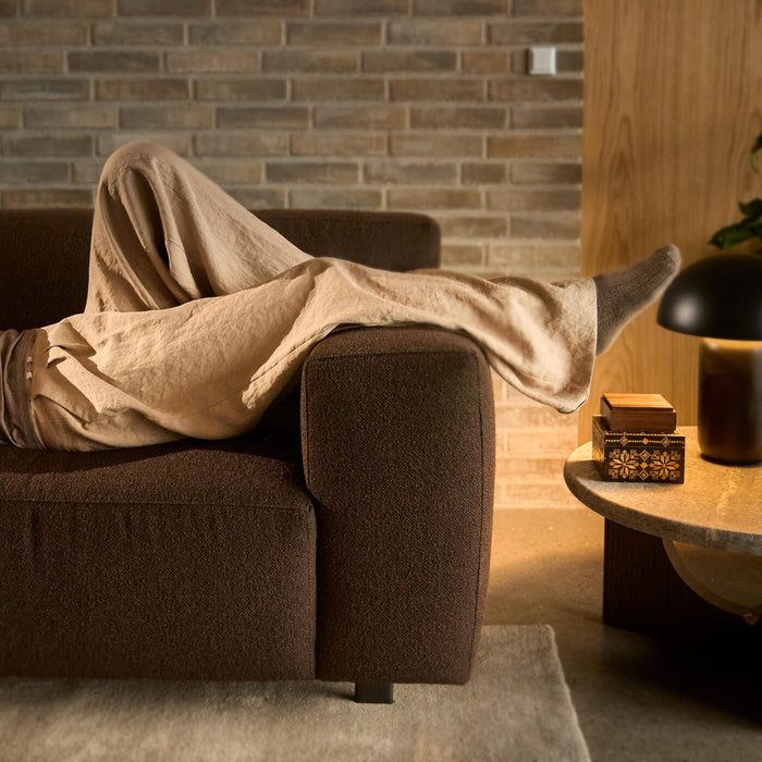 Ben der hviler på en mørkebrun sofa i en hyggelig stue med varm bordlampe, murstensvæg og en lille dekorativ æske på et rundt sofabord.