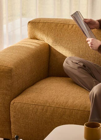 Person læser på en sennepsgul sofa i varmt dagslys, med et krus på bordet og lyse gardiner i baggrunden.