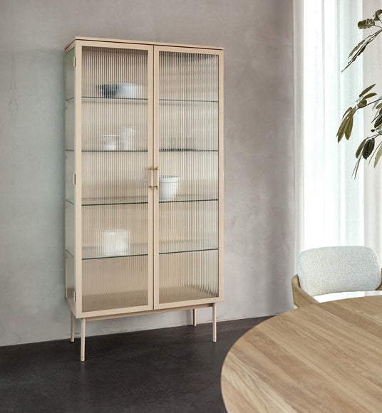 Beige vitrineskab med rillet glas på slanke ben i en lys spiseplads med rundt træbord, polstret stol og let gardin.