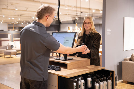Butiksmedarbejder i ILVA-trøje rækker en lille flaske til kunde ved skranke med skærm, der viser ILVAs hjemmeside i et møbelshowroom.