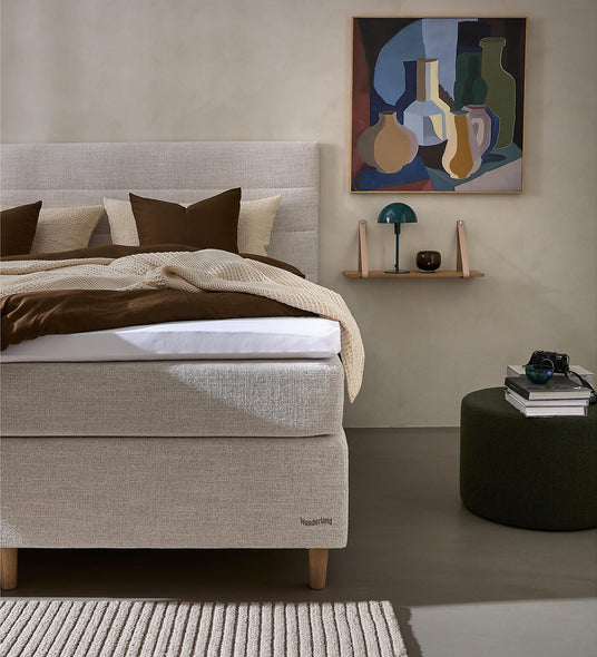 Minimalistisk soveværelsesscene med lys beige polstret seng, brune og cremefarvede puder og plaid, maleri og hylde med grøn lampe, mørkegrøn puff med bøger og tæppe; sengebunden er mærket Wonderland.