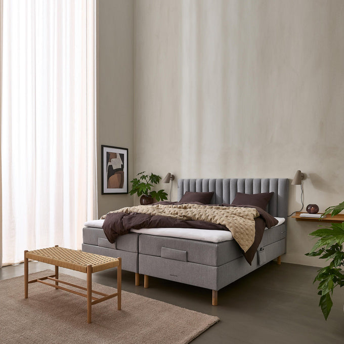 Lyst soveværelse med polstret grå seng, brunt sengetøj og quilt, planter, væglamper, naturfarvet tæppe og fletbænk for enden af sengen.
