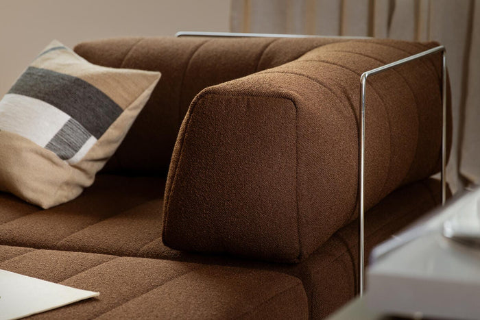 Nærbillede af brun modulsofa med metalarmlæn og pude i beige og grå ved et vindue med lyse gardiner.