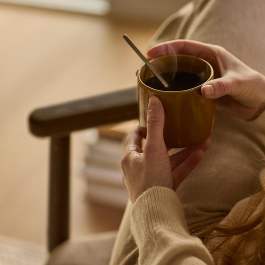 Nærbillede af hænder, der holder et varmt okkergult krus kaffe med ske, iført blød strik i en hyggelig stue.