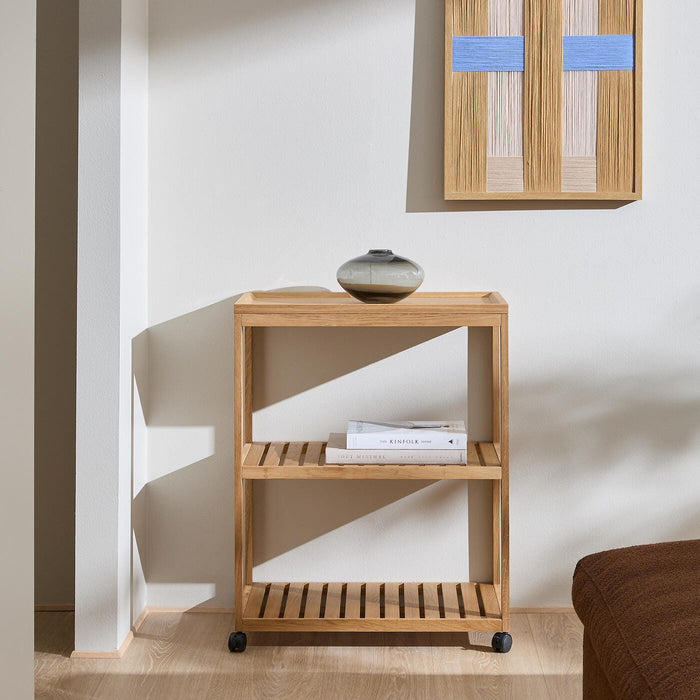 Minimalistisk reol i lyst egetræ på små hjul med lamelhylder; lille keramikvase ovenpå, vægbillede med blå detaljer og bøgerne Soft Minimal og The Kinfolk Home på midterhylden i en lys stue med sollys.