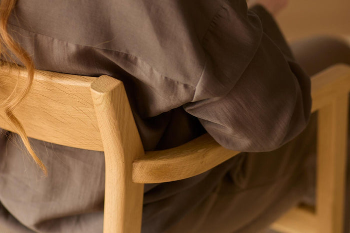 Nærbillede bagfra af en person, der sidder på en lys træstol og holder et noteshæfte i varme, beige nuancer.