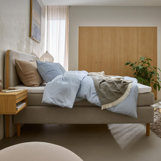Lyst og roligt soveværelse med seng med lyseblåt sengetøj, beige puder og gråt plaid, træ-natbord og garderobeskabe samt grøn plante i blødt dagslys.