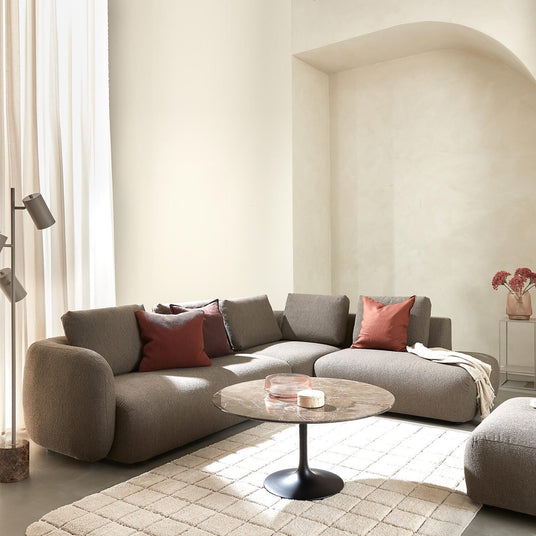 Lyst, minimalistisk stue med grå modulsofa, terrakotta puder, rundt marmorbord med sort fod, lyse gardiner og en niche med vase og blomster på et sidebord.