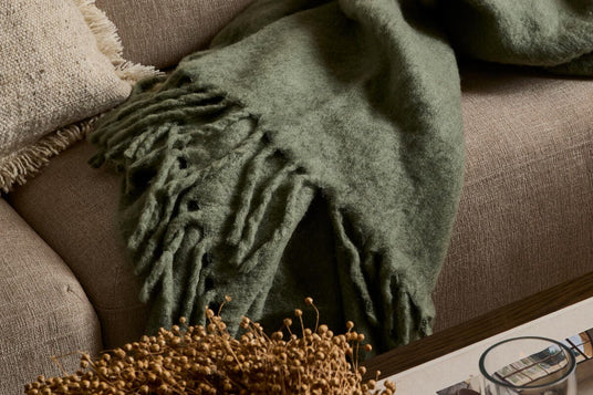 Nærbillede af et blødt grønt plaid med frynser lagt over en beige sofa med en tekstureret pude; på sofabordet står et stearinlys i glas og et magasin.