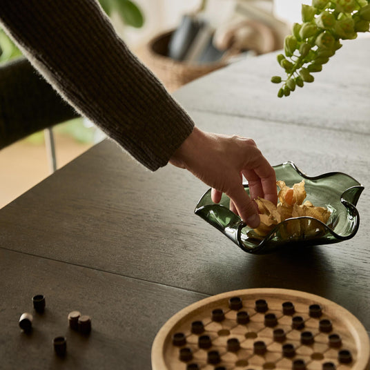 Hånd tager snacks fra grøn bølget glasskål på mørkt spisebord, med brætspil og planter i baggrunden.