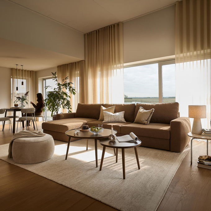 Lyst stuebillede med beige gardiner, stor brun hjørnesofa, to runde sofaborde med lys og skål, puf og spiseafdeling i baggrunden med plante og person i silhuet; store vinduer lukker dagslys ind.
