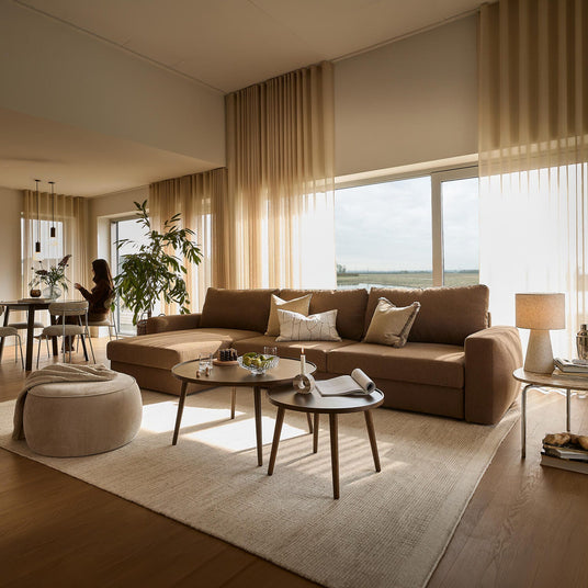 Lyst stuebillede med beige gardiner, stor brun hjørnesofa, to runde sofaborde med lys og skål, puf og spiseafdeling i baggrunden med plante og person i silhuet; store vinduer lukker dagslys ind.