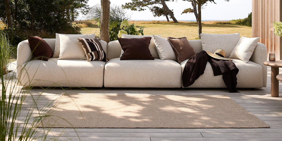 Beige modulsofa på træterrasse udendørs med brune og lyse puder, tæppe og sidebord i træernes skygge med udsigt mod eng og vand.