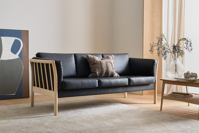 Sort lædersofa med lys træramme i en minimalistisk stue med beige tæppe, sofabord med glasvase og grene, vægkunst og lyse gardiner.
