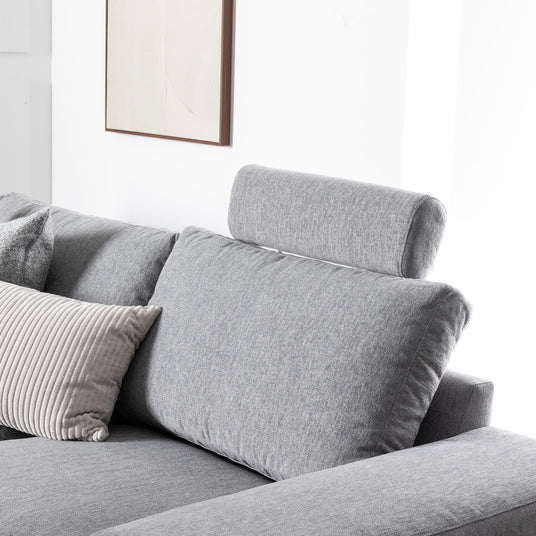 Grå stofsofa med justerbar nakkestøtte og beige/grå pyntepuder mod hvid væg med enkel ramme i et lyst, minimalistisk stuemiljø.