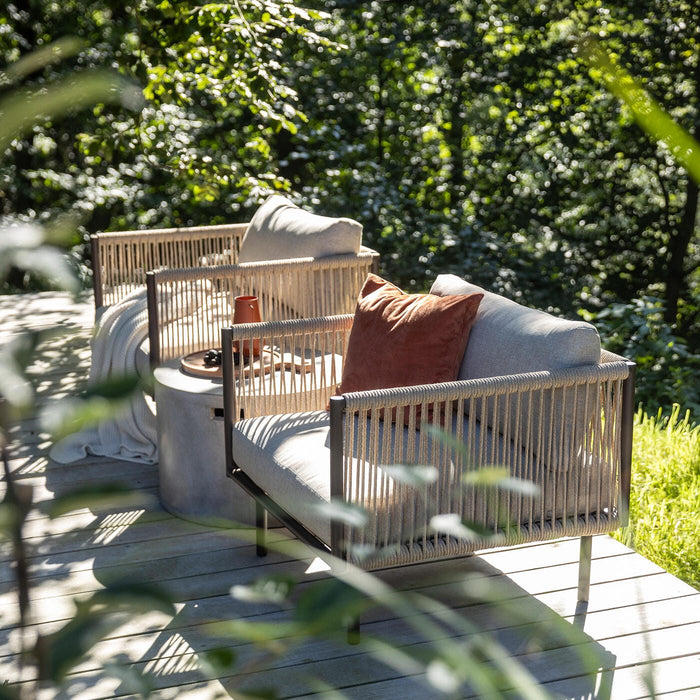 To moderne udendørs lænestole med rebdetaljer på træterrasse, frodig skov i baggrunden, puder og plaid samt bakke med kop i sollys.
