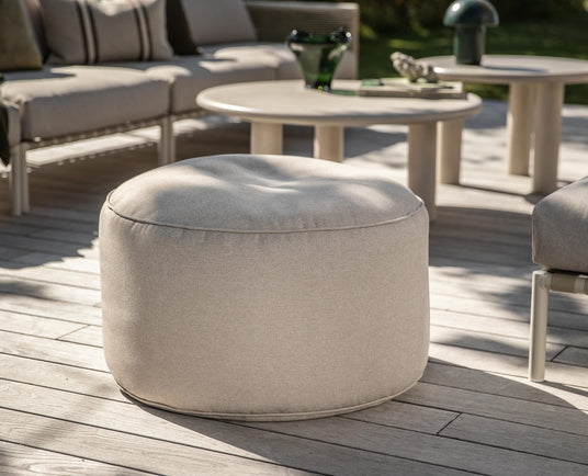 Beige rund udendørs puf på træterrasse, omgivet af lyse loungemøbler og sofaborde i dagslys.