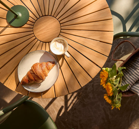 Solbelyst rundt træbord med en croissant på tallerken og cappuccino; grøn stol og solsikker i kurv på terrasse.