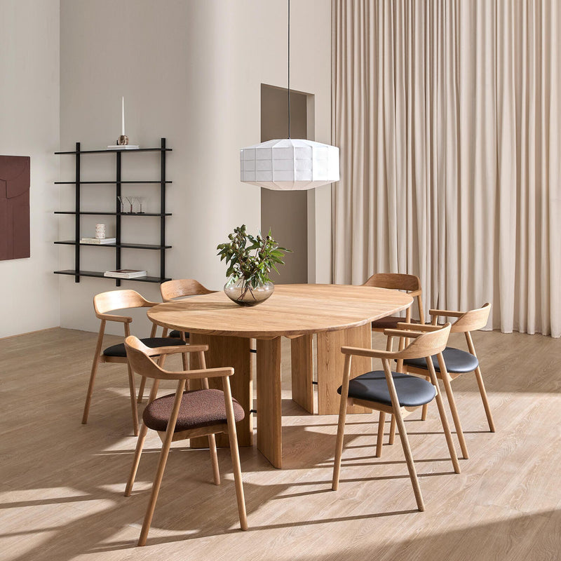 Minimalistisk spisestue med rundt egetræsbord, seks træstole, hvid pendel og glasvase med grønne grene; beige gardiner og sort hylde i baggrunden.