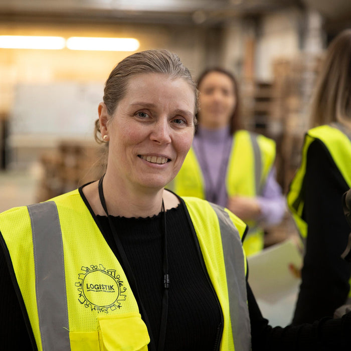 Person i gul sikkerhedsvest med LOGISTIK‑logo i lager-/produktionsmiljø, kolleger i baggrunden.
