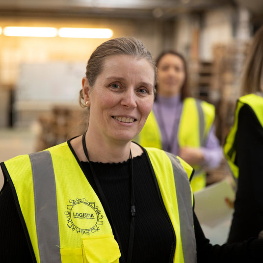 Person i gul sikkerhedsvest med LOGISTIK‑logo i lager-/produktionsmiljø, kolleger i baggrunden.