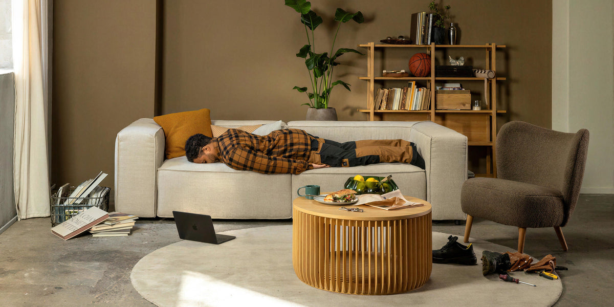 Stue med beige sofa hvor en person hviler, rundt tæppe og træ-sofabord, reol med bøger og planter, blødt dagslys fra et vindue.