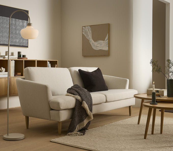 Lysebeige sofa med mørk pude og plaid, gulvlampe, runde sofaborde med lys og vase i et neutralt skandinavisk stue-miljø med vægkunst og lyst træ.