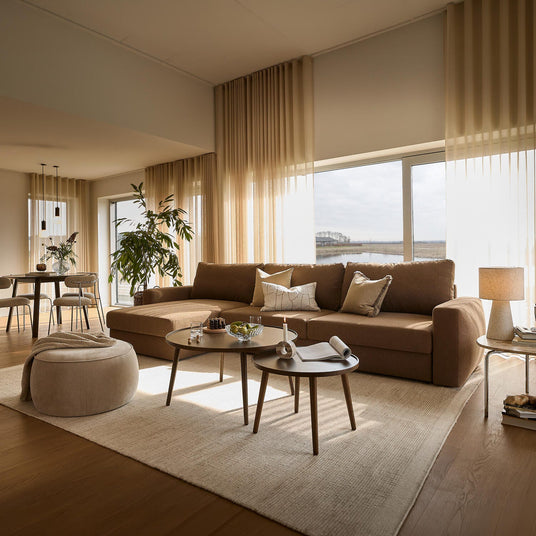Lyst og varmt stueinteriør med beige gardiner, brun hjørnesofa, to runde sofaborde, puf, planter, spiseplads og store vinduer mod vandet.