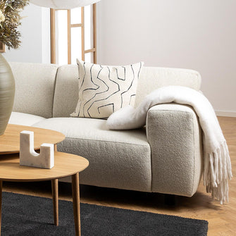 Lys skandinavisk stue med beige hjørnesofa, runde sofaborde i egetræ på sort tæppe og to store rispendler; stor vase med tørrede planter og grafisk mønstret pude.