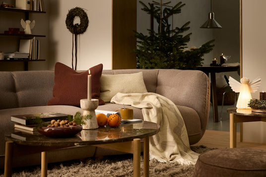 Stue med brun sofa, juletræ, tændte lys, puder og plaid på sofaen; varm og hyggelig julestemning.