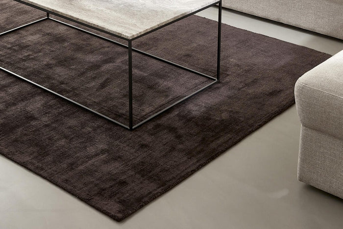 Beige sofa og lænestol med mørkebrun tæppe og rektangulært sofabord med stenplade og sort metalstel; vase med tørrede grene, lille lyslygte og en opslået bog på bordet.