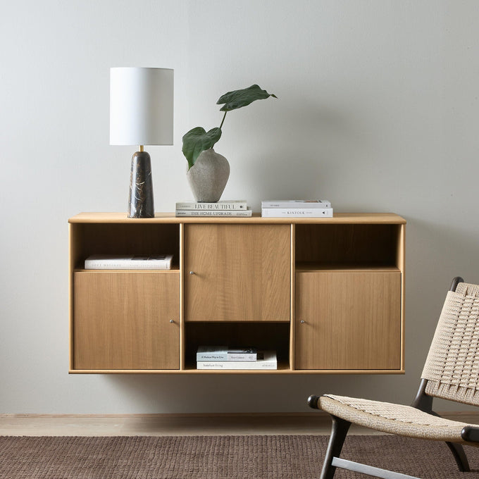 Væghængt skænk i lyst træ set forfra med bordlampe, stenvase med grøn bladplante og stabler af bøger – LIVE BEAUTIFUL, THE HOME UPGRADE og THE KINFOLK – ved siden af en fletlænestol i en lys stue.