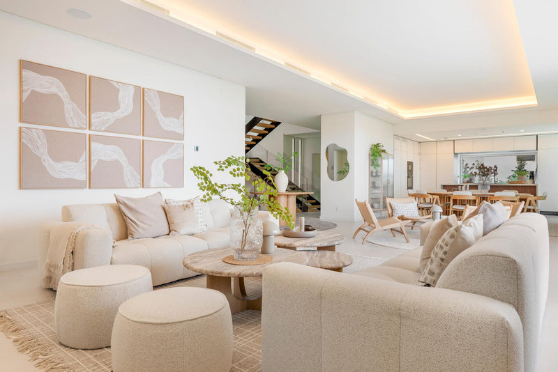 Lys, åben planløsning med beige sofa, runde sofaborde i træ, seksdelt vægkunst og grønne planter; skandinavisk stue med spisestue i neutrale toner.