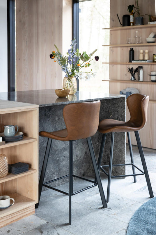 Lyst, moderne køkkenø i mørk sten med to brune barstole og vase med friske blomster.